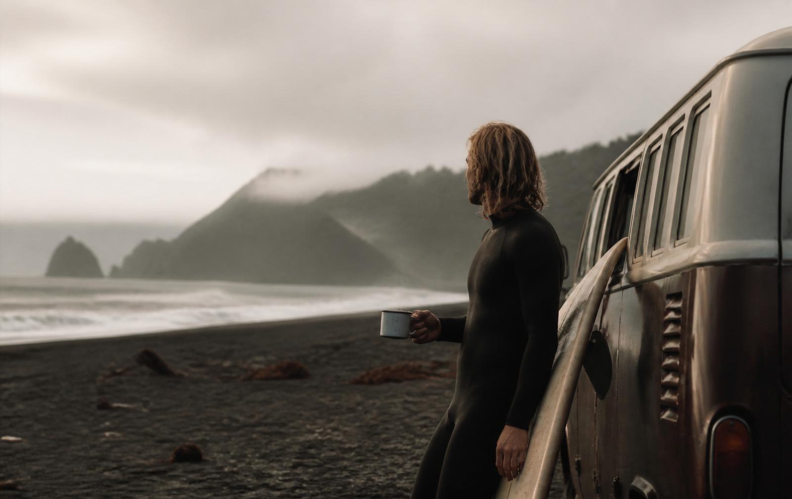 Surfer holding an enamel mug beside a van at a black sand beach at sunrise